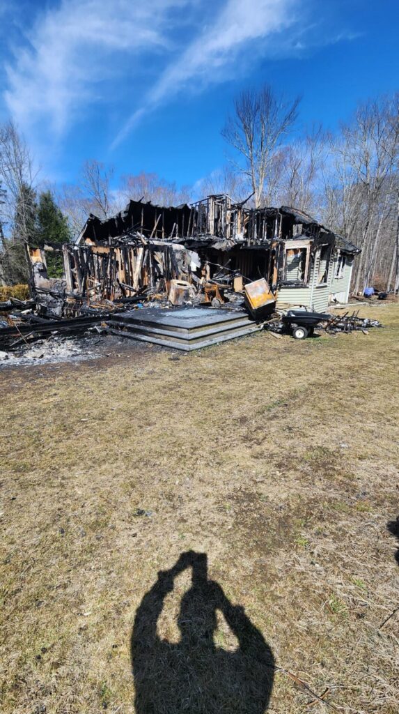 A fire-damaged house stands heavily charred with a collapsed roof, exposed framing, and debris scattered across the yard