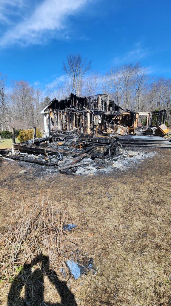 A severely burned house stands amid ashes and debris under a clear blue sky, with charred framing, collapsed remains, and scorched ground.
