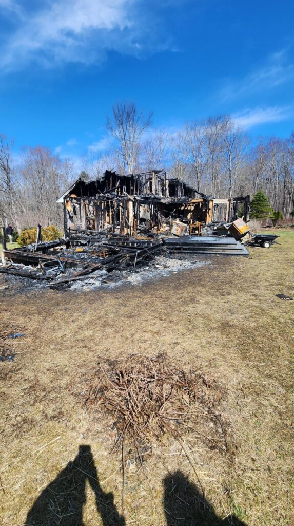 A badly burned house stands amid a scorched yard, with charred beams, collapsed debris, and leafless trees