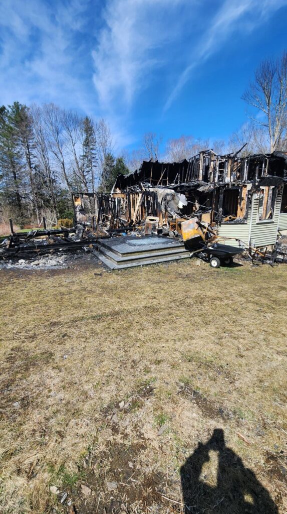A charred house stands after a severe fire, with collapsed walls, burned debris, and scorched siding