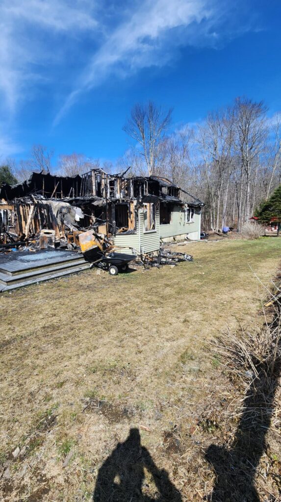 Severely burned house exterior with collapsed roof, charred siding, and debris scattered across the yard under a clear blue sky after a fire.