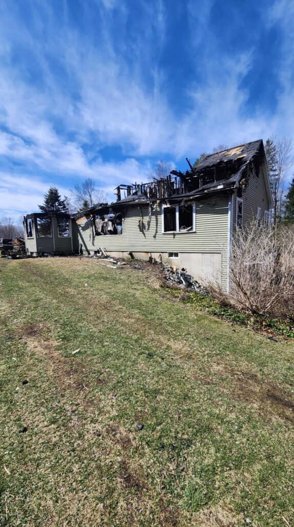 A fire-damaged house stands with a charred roof, broken windows, and exposed framing beside a grassy yard under a bright blue sky after the blaze.