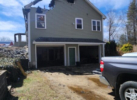 Severe fire damage leaves a charred New Braintree, MA home with a burned roofline, dark garage openings, and a driveway beside a pickup truck.