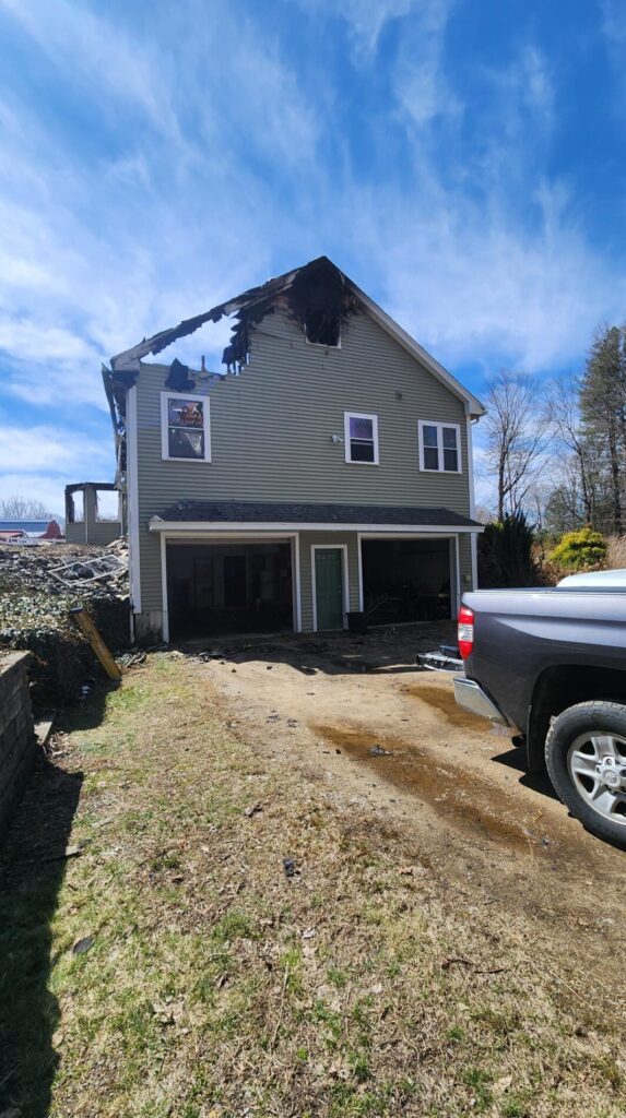 Severe fire damage leaves a charred New Braintree, MA home with a burned roofline, dark garage openings, and a driveway beside a pickup truck.