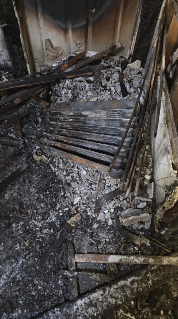 Severe fire damage leaves charred wall framing and ash-covered debris scattered across the damaged interior room.