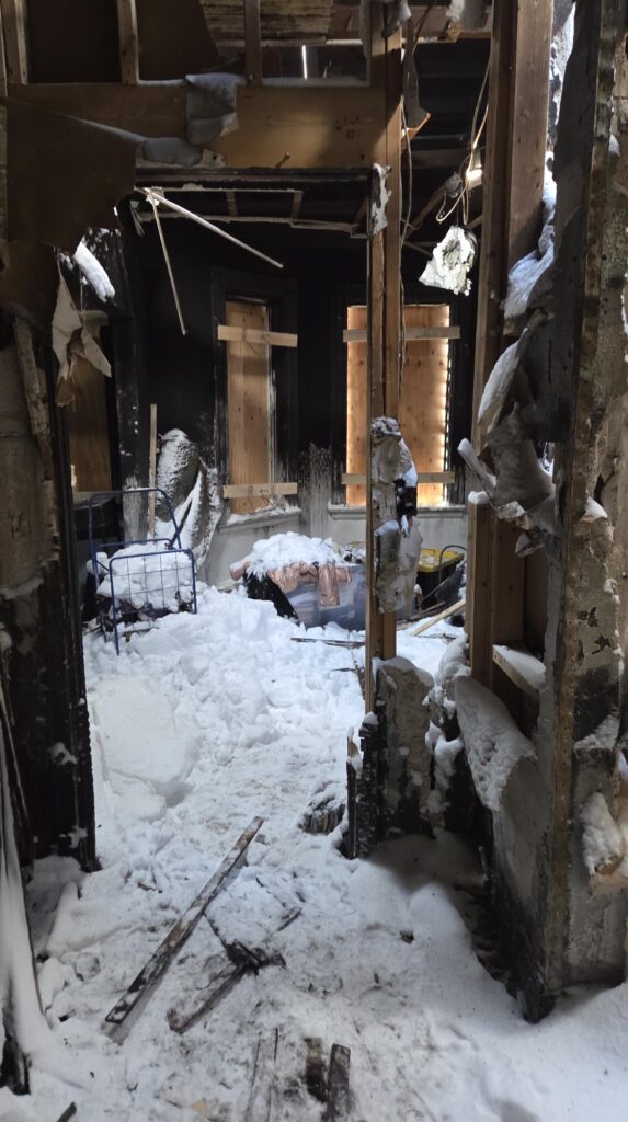 Fire-damaged interior of a gutted home filled with snow, charred walls, exposed framing, and boarded windows