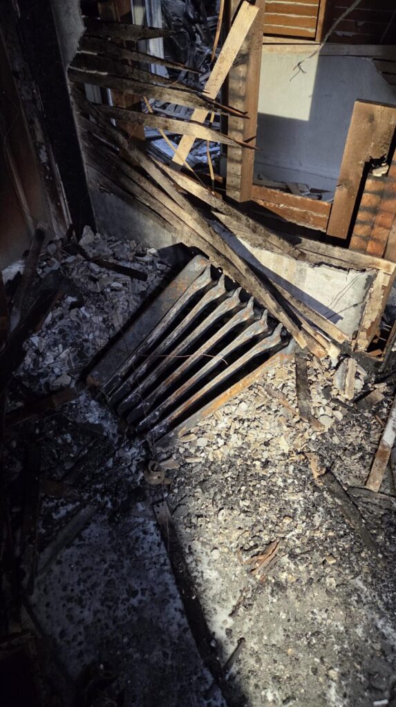 Severe fire damage leaves charred wall framing and ash-covered debris scattered across the damaged interior room.