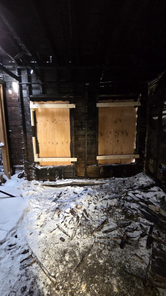 Severely fire-damaged interior with boarded-up windows, blackened walls, and snow-covered debris scattered across the floor