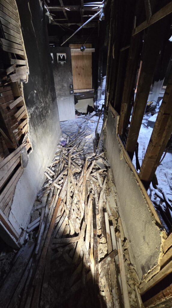 Fire-damaged hallway with exposed studs, blackened walls, scattered debris, and a boarded doorway inside a heavily damaged home after a severe blaze.