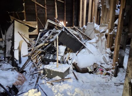 Collapsed interior framing and scattered debris fill a snow-covered room after severe structural damage, with exposed beams and a partly torn roof.