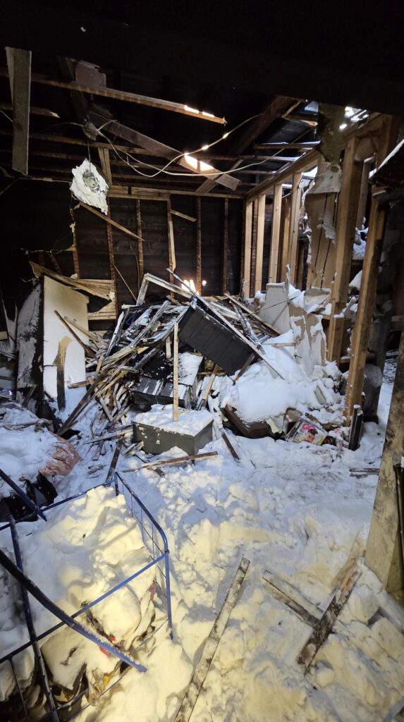 Collapsed interior framing and scattered debris fill a snow-covered room after severe structural damage, with exposed beams and a partly torn roof.