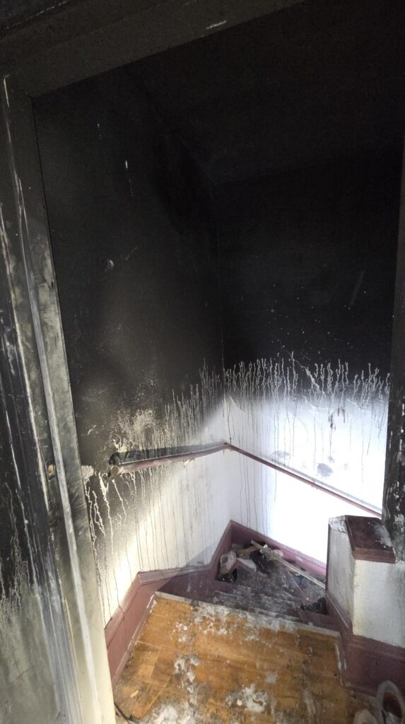 A heavily fire-damaged stairwell shows black soot on the walls and ceiling, smoke streaks down the painted surfaces, and debris on the steps.
