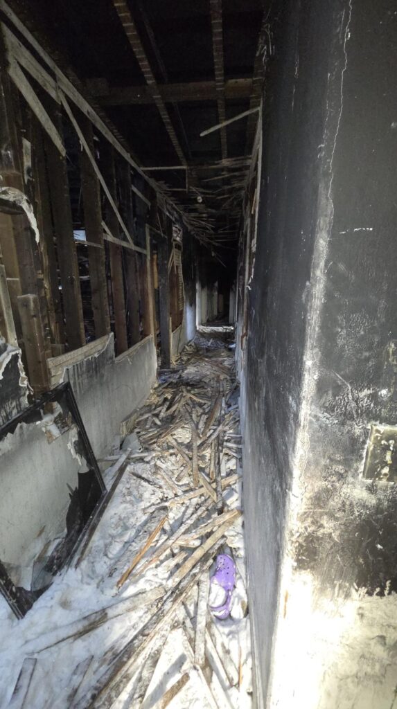 A charred hallway with exposed framing, fallen debris, and smoke-stained walls shows severe fire damage and destruction along the narrow passage.