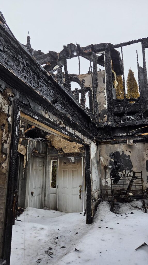 Inside a burned house, charred framing, open roof beams, and snow covering the floor show severe fire damage