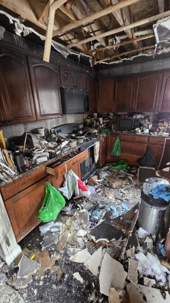 Fire-damaged kitchen with burned ceiling joists, smoke-stained cabinets, debris-covered floor, and damaged appliances