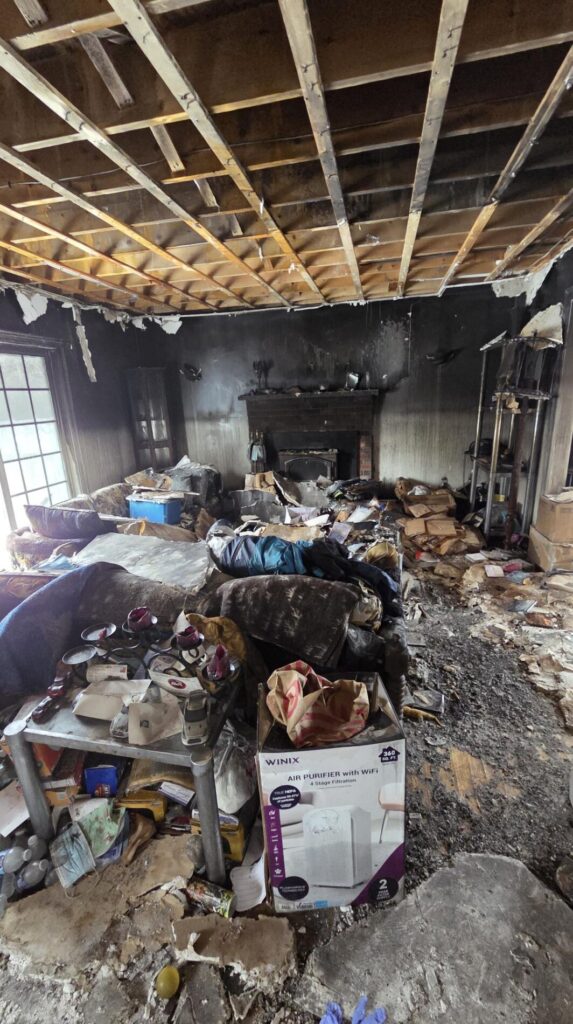 Severe fire damage fills a living room with a burned ceiling, soot-stained walls, scattered debris, and ruined furniture