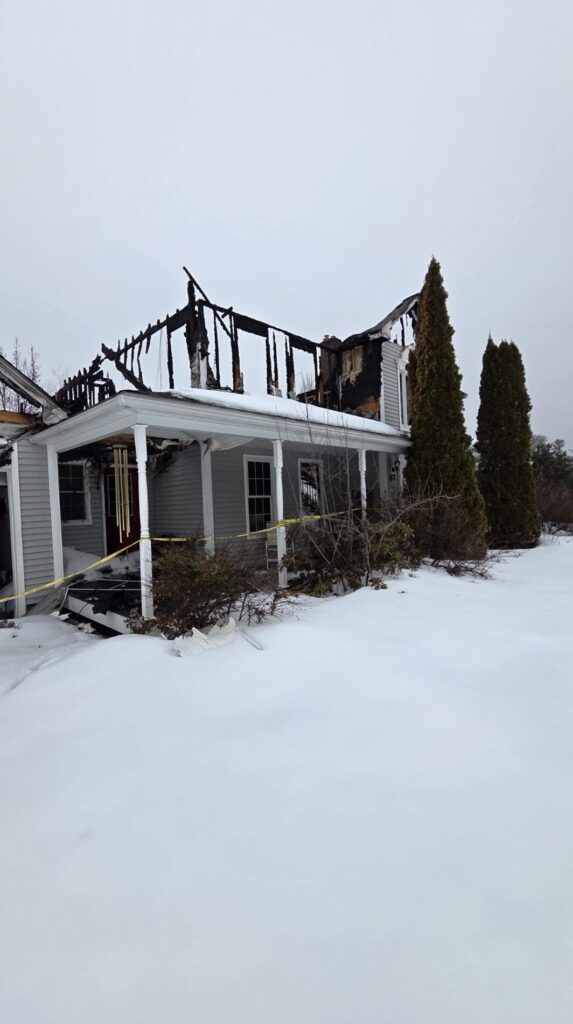 A heavily fire-damaged house stands in a snowy yard with a burned roof, charred framing, and caution tape across the front porch