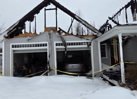 Snow covers a fire-damaged home in Hardwick, MA, with a burned garage, exposed roof trusses, and yellow caution tape across the driveway below.