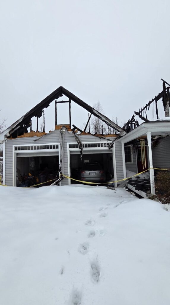 Snow covers a fire-damaged home in Hardwick, MA, with a burned garage, exposed roof trusses, and yellow caution tape across the driveway below.