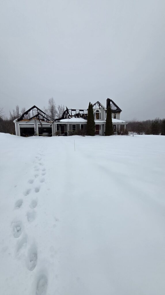 A fire-damaged house stands in a snowy yard with footprints leading toward it under a gray winter sky, with charred roof framing and bare trees.