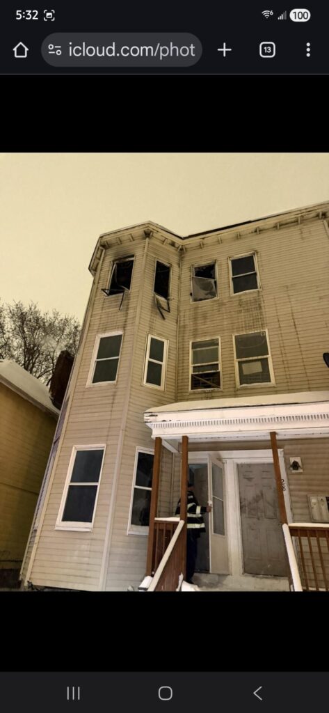 A beige three-story home shows shattered upper windows and winter snow on the porch, while a person stands near the entryway after fire damage.