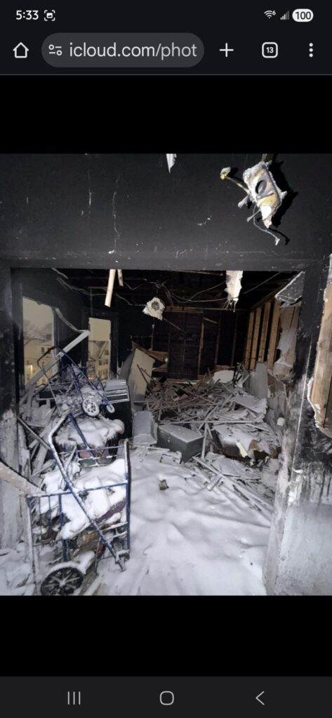 A fire-damaged room with blackened walls, collapsed debris, and snow-covered wreckage fills the space after severe structural damage.