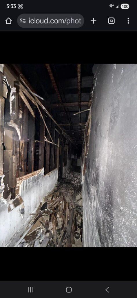 A fire-damaged hallway with blackened walls, exposed framing, and debris strewn across the floor inside a severely burned building.