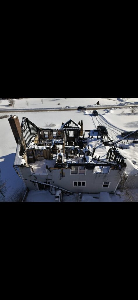 Aerial view of a fire-damaged house with a snow-covered roof, charred upper framing, and exposed rooms